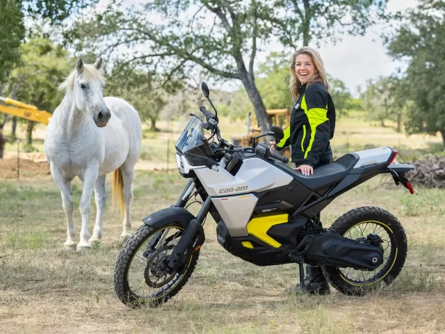 A smiling woman sits on a Can-Am Pulse or Origin electric motorcycle, with a white horse in the background in a natural setting.