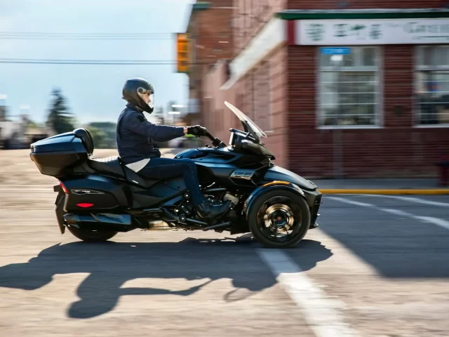 A helmeted biker drives a black trike through a city street, with a brick building in the background.